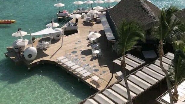 Un complexe hôtelier en bord de mer doté d’une terrasse en bois, de chaises longues, de parasols et d’une structure à toit de chaume.