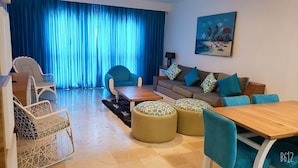 Un salon comprenant un canapé, une table basse, des poufs et des fauteuils. Un tableau est accroché au mur, et une fenêtre est ornée de rideaux bleus.