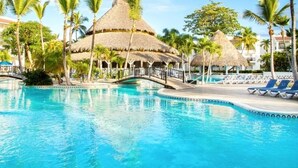 Une piscine d’hôtel agrémentée d’une structure à toit de chaume, de palmiers et de chaises longues.