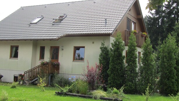 Une maison à deux étages avec un toit en pente, un balcon au deuxième étage et un petit jardin à l’avant.