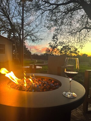 A round outdoor table with a fire pit in the center, a glass of red wine, and a view of trees and a sunset in the background.
