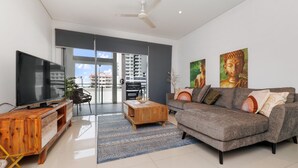 A modern living room with a flat-screen TV, a wooden coffee table, a grey sofa with cushions, and two Buddha statues. - Stylish 3 BR Apartment in The Heart of Darwin CBD with Pool (Darwin City)