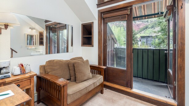 Een kamer met een houten bank, een bureau met een koffiezetapparaat, een grote spiegel en een glazen schuifdeur naar een balkon.
