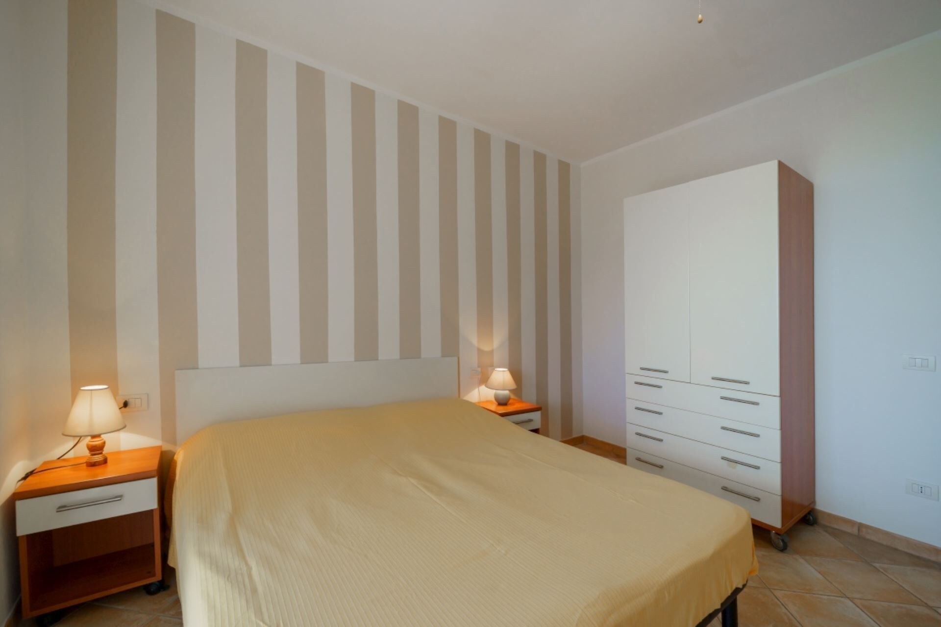 A bedroom with a bed, a nightstand with a lamp, a wardrobe, and a striped wall.