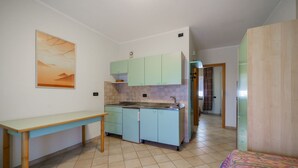 Cuina compacta amb armaris verdosos, una taula de dinar petita i un llit amb llençols de colors.