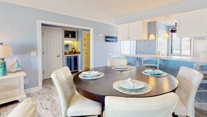 A dining area with a round table set for four, surrounded by white chairs. There is a kitchen visible in the background with white cabinets and a yellow door. - Oceanfront Top Floor Ocean Dunes, Bunk Room, Indoor/Outdoor Pools, Hot Tub, Tennis (Kure Beach)