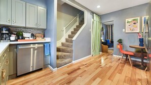 Eine moderne Küche mit grauen Schränken, einem Geschirrspüler und einer Kaffeemaschine. Eine Treppe führt zu einem Wohnbereich mit einer Couch und einem Esstisch.