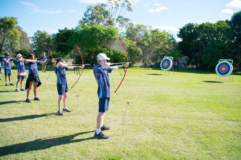 A group of people practicing archery outdoors on a grassy field with trees in the background.