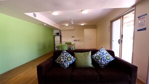 A living room with a brown sofa, patterned cushions, a green wall, and a kitchen in the background. - Gold Coast Performance Centre - Hostel (Runaway Bay)