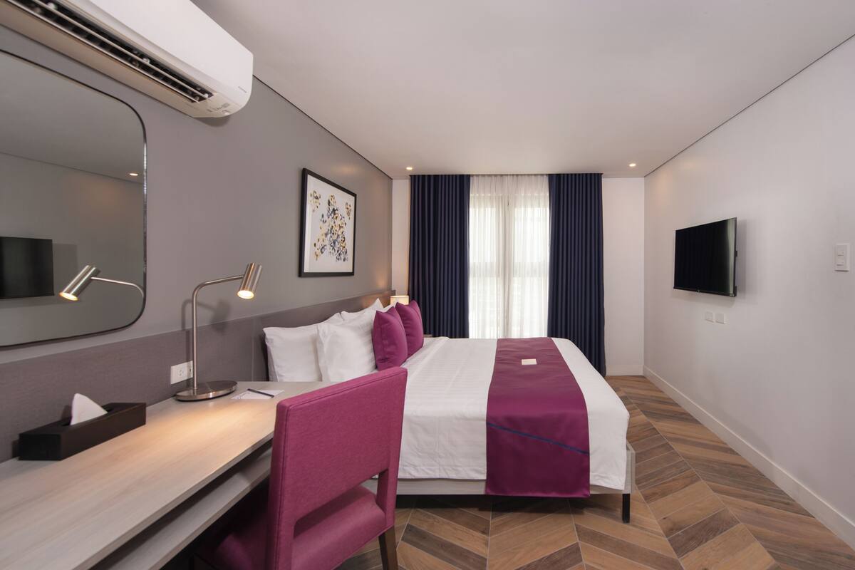 Habitación de hotel con cama, mesita de noche, lámpara, silla, televisor y ventana con cortinas.