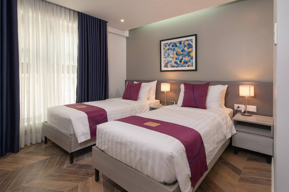 Habitación de hotel con dos camas, cada una con cubrecama blanco y detalles morados, una mesita de noche con lámpara, un cuadro en la pared y un ventanal con cortinas translúcidas.