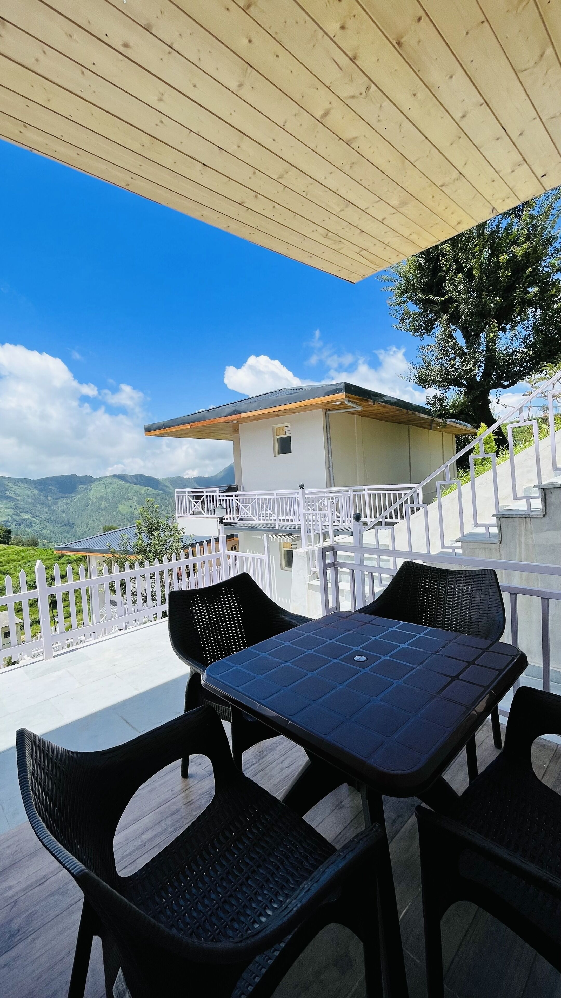 An outdoor patio with a table and chairs, overlooking a mountainous landscape and a white building with a balcony.