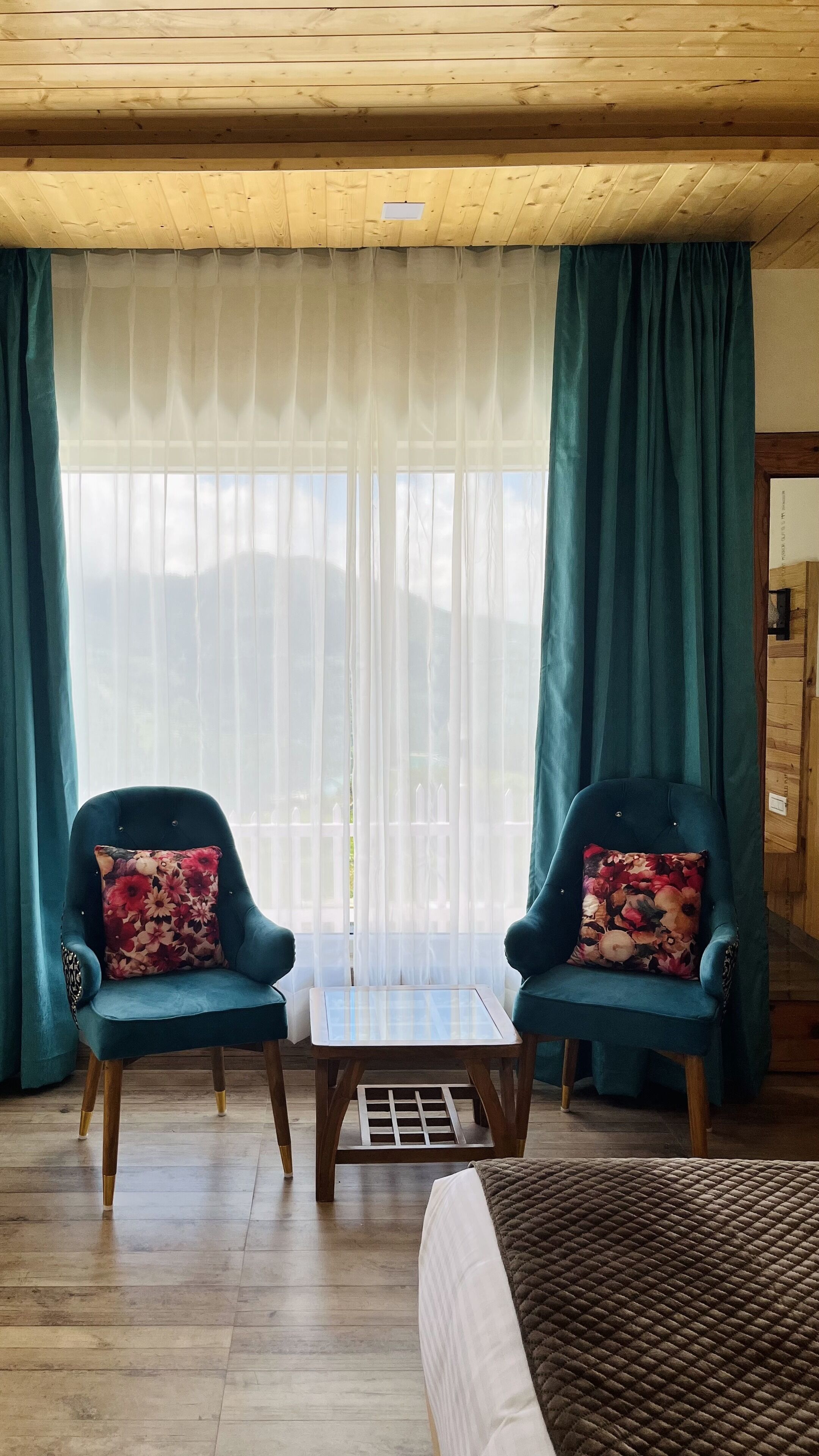 A room with two teal armchairs, a small wooden table, and a bed with a brown quilt.