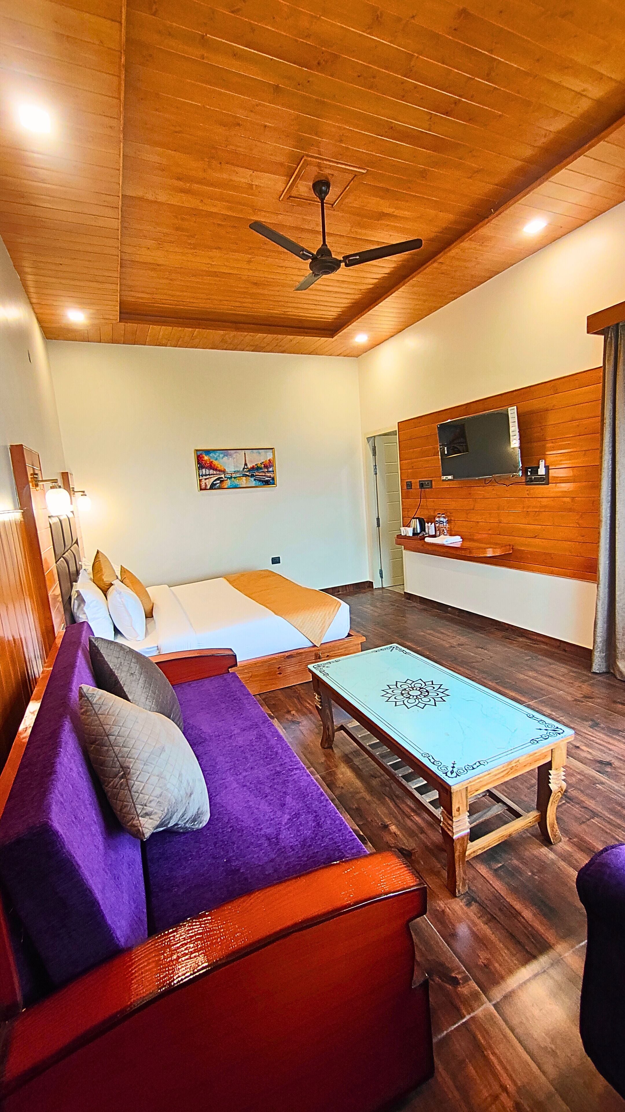 A room with a wooden ceiling, a bed with a white and orange bedspread, a purple sofa with cushions, a wooden coffee table with a blue patterned top, and a television mounted on the wall.