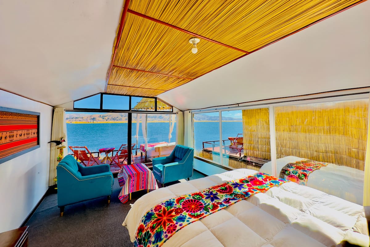 A room with a bed, two blue chairs, a small table, and a view of the sea.