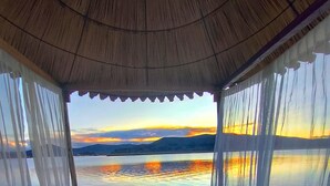 A bed with a colorful blanket and pillows, a view of a sunset over water, and a thatched roof structure.