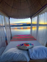 Una cama con una manta y almohadas coloridas, vista de un atardecer sobre el agua y una estructura de techo de paja.
