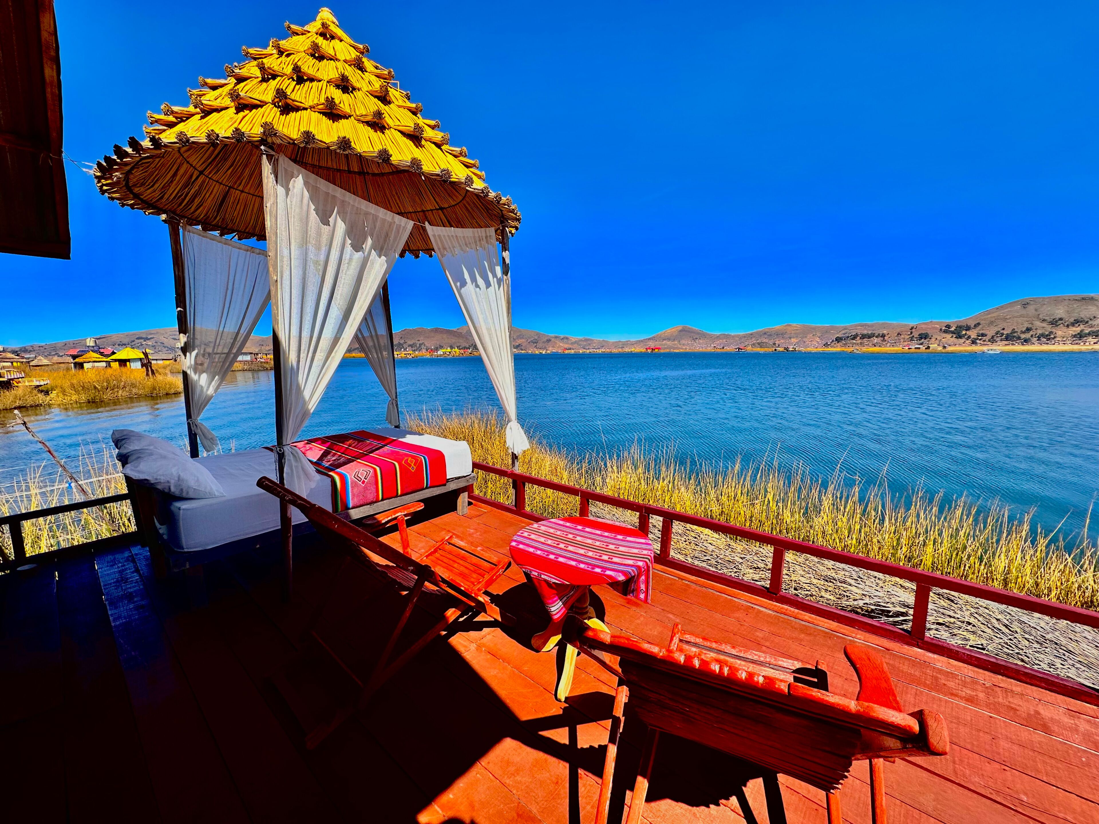 A wooden deck with a canopy and two beds, overlooking a lake with distant hills.