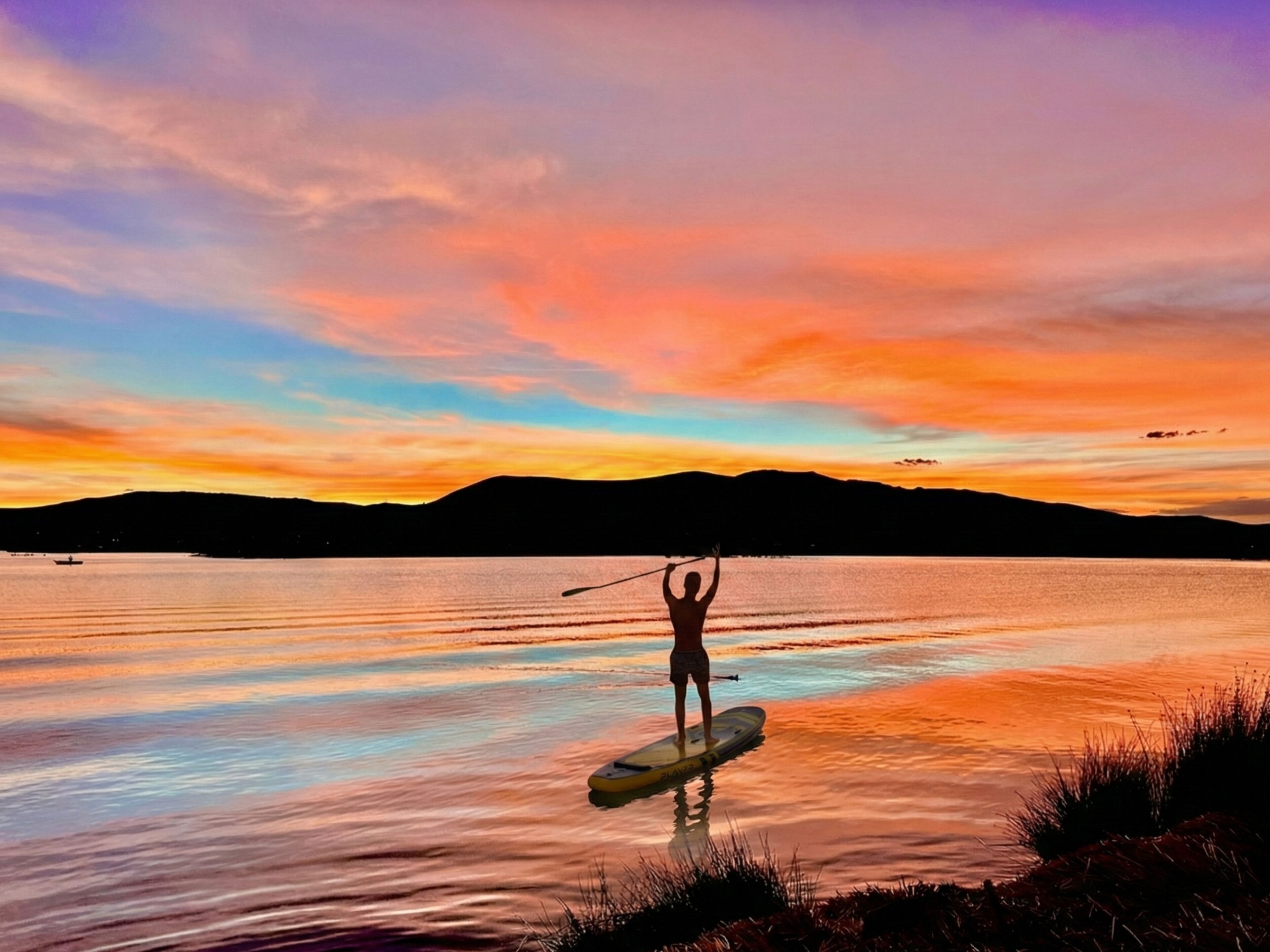 A person paddleboarding on a calm lake during sunset with mountains in the background.