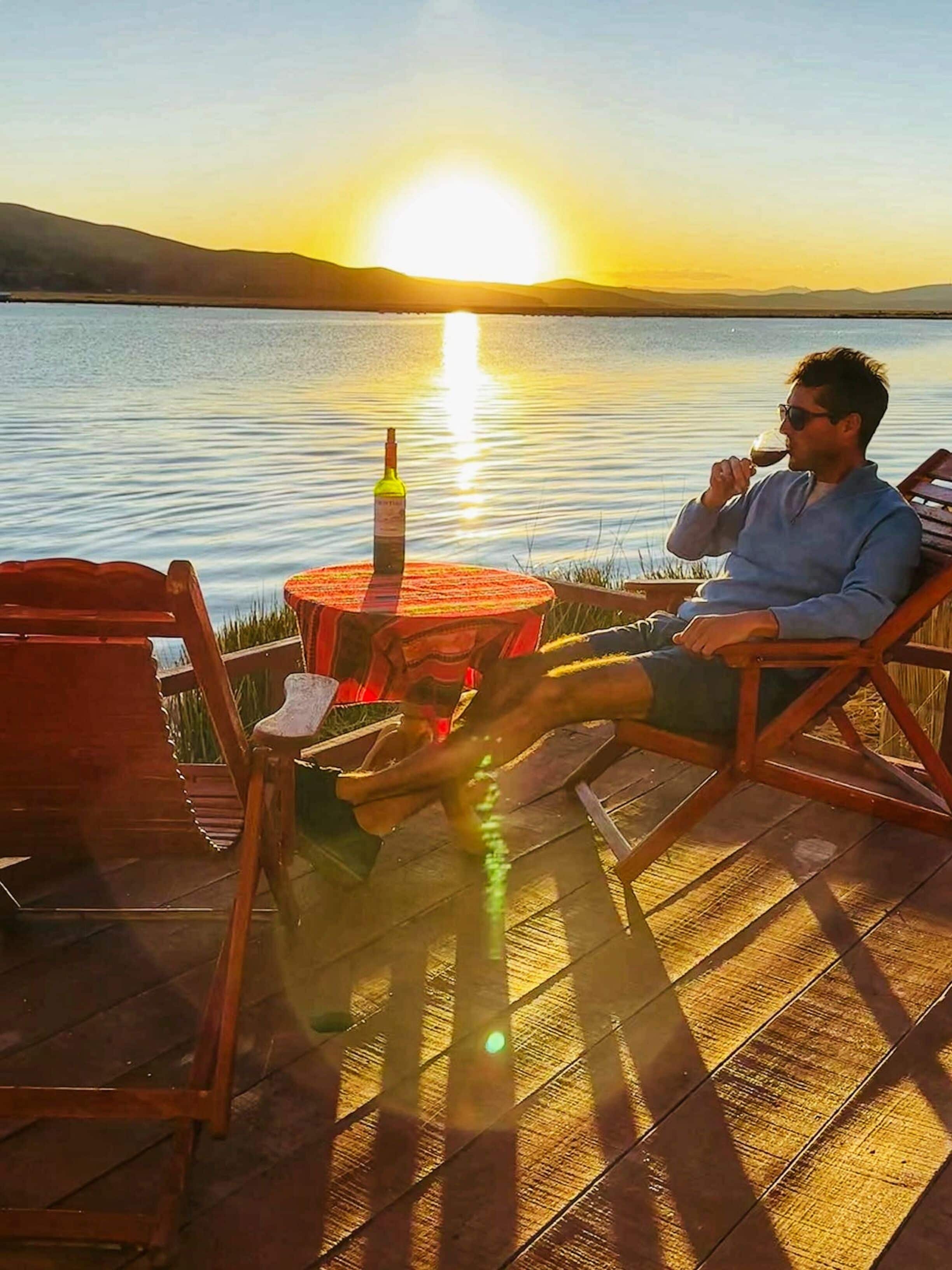 A person relaxing on a wooden deck by a lake at sunset, with a bottle of wine and a chair.