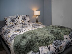 Un dormitorio con cama, ropa de cama floral, un plaid suave color verde, una lámpara de noche y un armario.