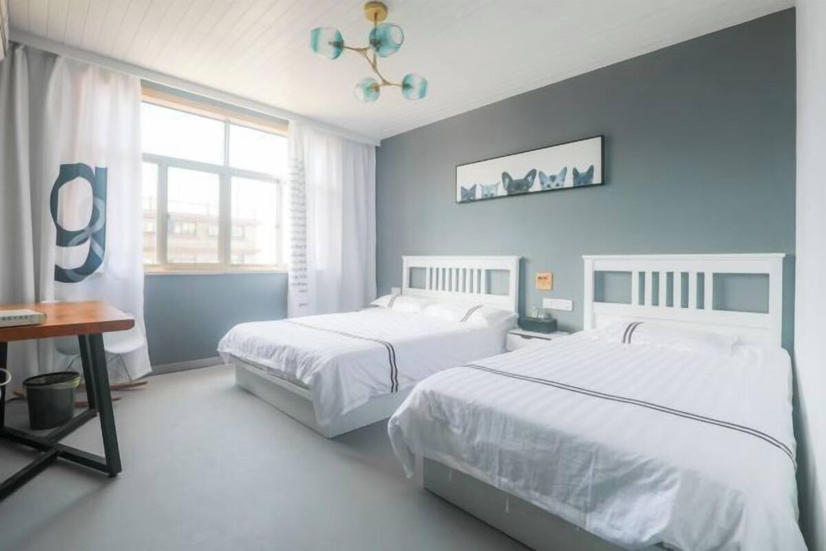 A room with two beds, a desk, a chair, a radiator, and a window with curtains.