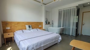 A modern hotel room with a wooden headboard, a large bed, a white bathtub, and a small wooden bench.
