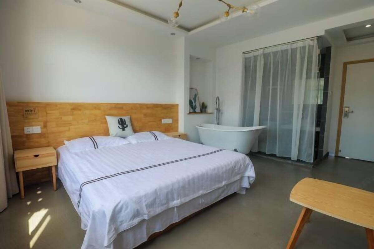 A modern hotel room with a wooden headboard, a large bed, a white bathtub, and a small wooden bench.
