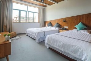 Quarto de hotel com duas camas, uma cabeceira de madeira, uma janela com cortinas, uma mesinha com uma planta e um tapete redondo.