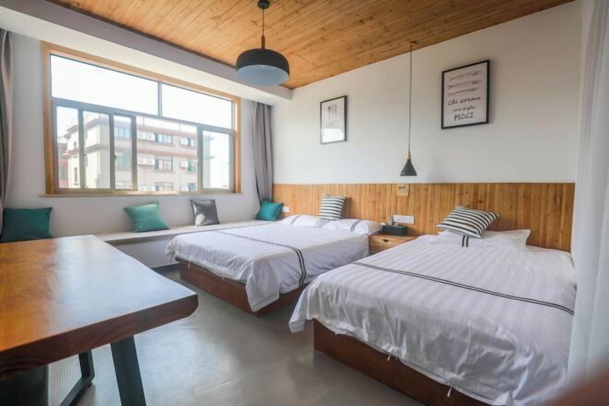 Quarto de hotel com duas camas, teto de madeira, uma janela grande e uma mesa.