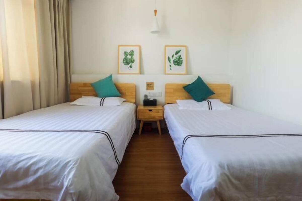 A hotel room with two single beds, wooden headboards, white bedding, teal pillows, a nightstand with a lamp, and framed leaf artwork on the wall.