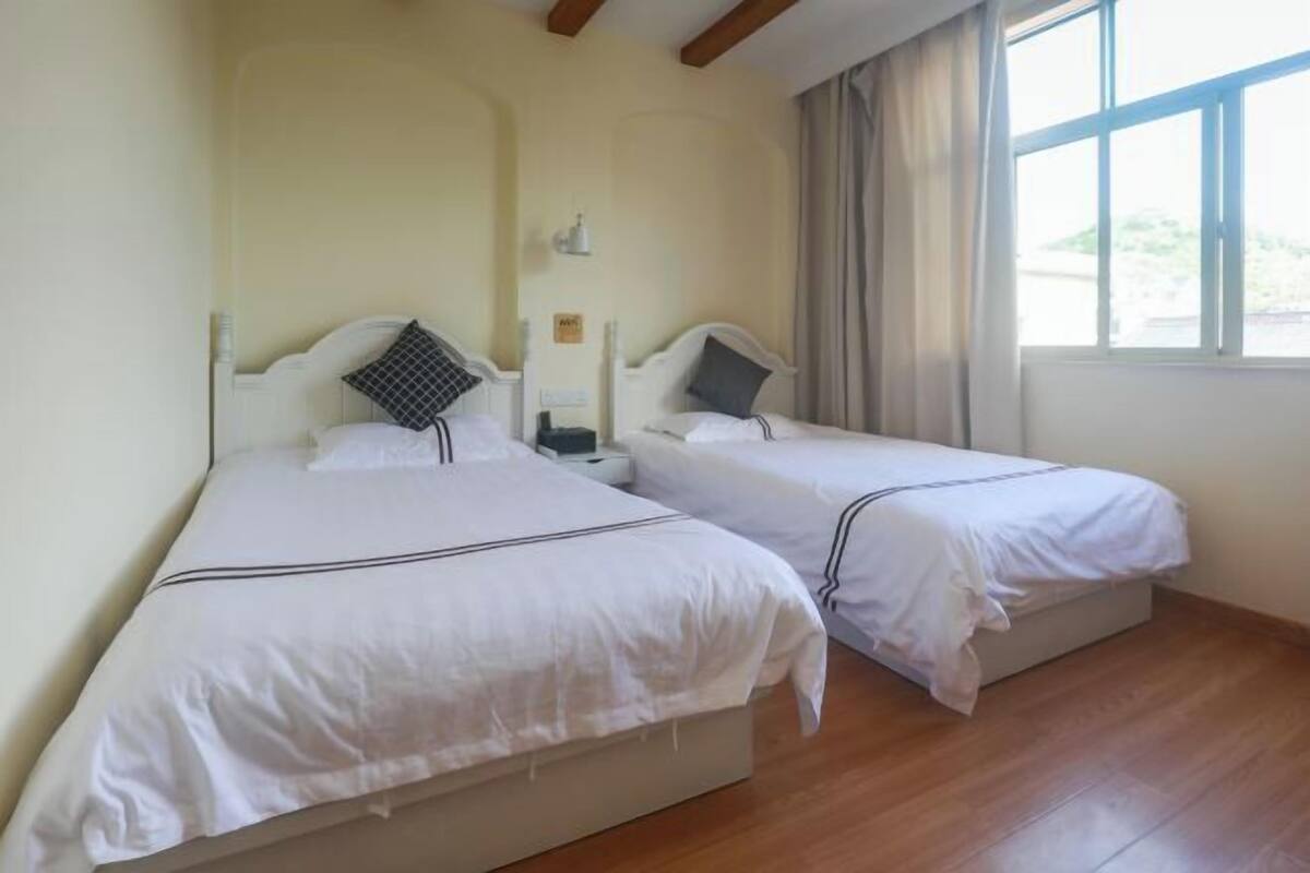 A hotel room with two single beds, wooden flooring, a window with curtains, and a small bedside table with a lamp.