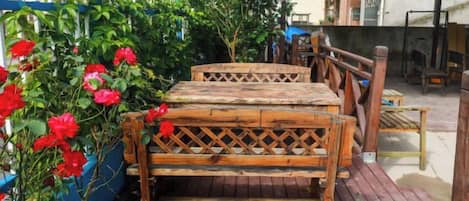 A wooden bench with a lattice backrest, positioned on a wooden deck with a blue planter of red flowers to the left.