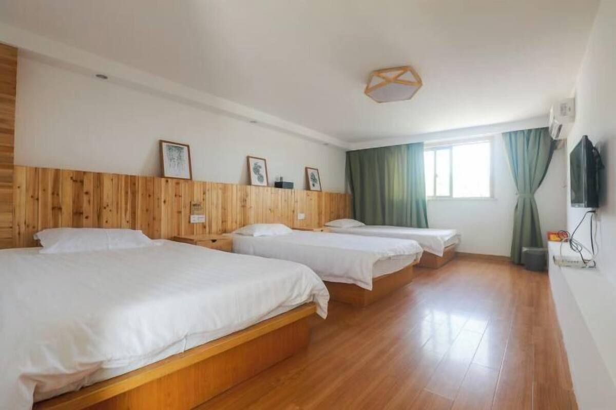 A hotel room with three wooden beds, a large window with green curtains, and a wooden ceiling light fixture.