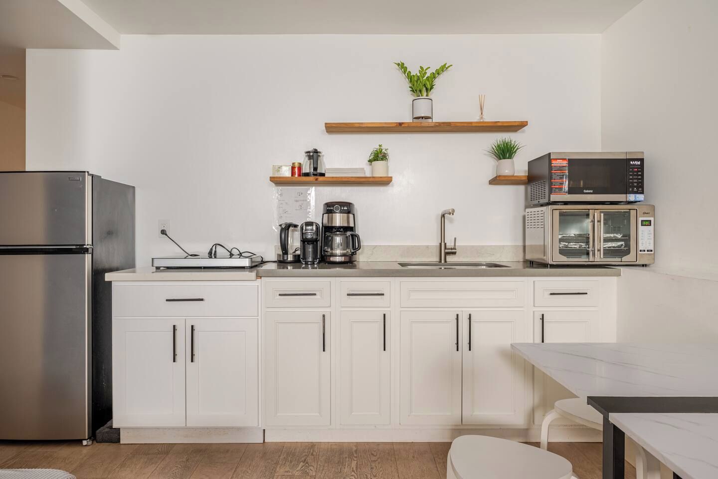 Eine moderne Küche mit weißen Schränken, einem Edelstahlkühlschrank, einer Kaffeemaschine, einer Mikrowelle und Holzregalen mit Pflanzen.