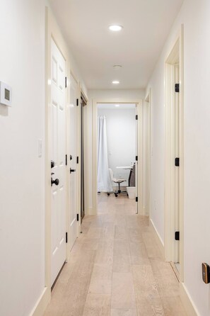 Un pasillo con puertas blancas, una zona de ducha y piso de madera de tono claro.