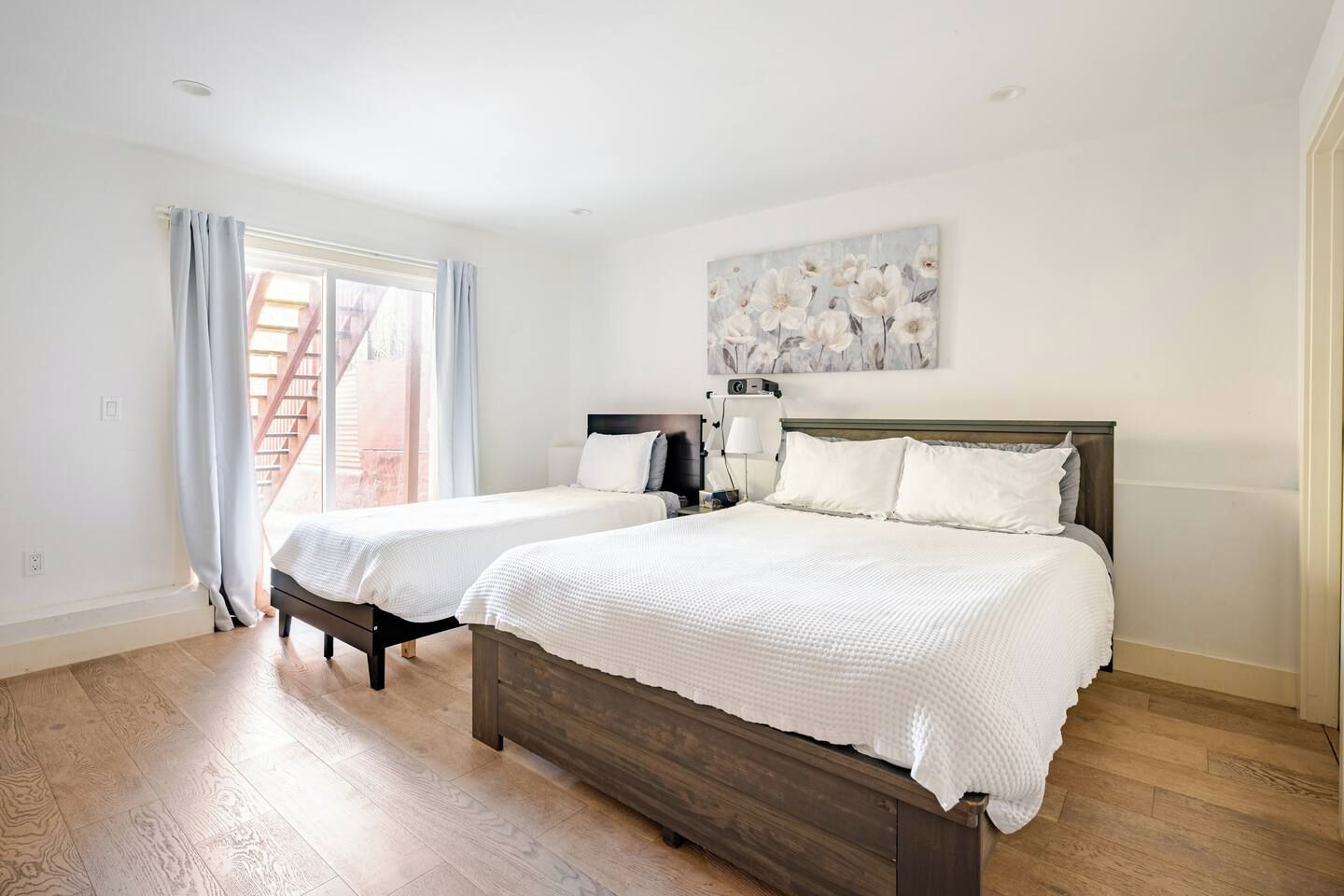 A bedroom with two beds, a window with curtains, a painting on the wall, and a staircase visible outside.