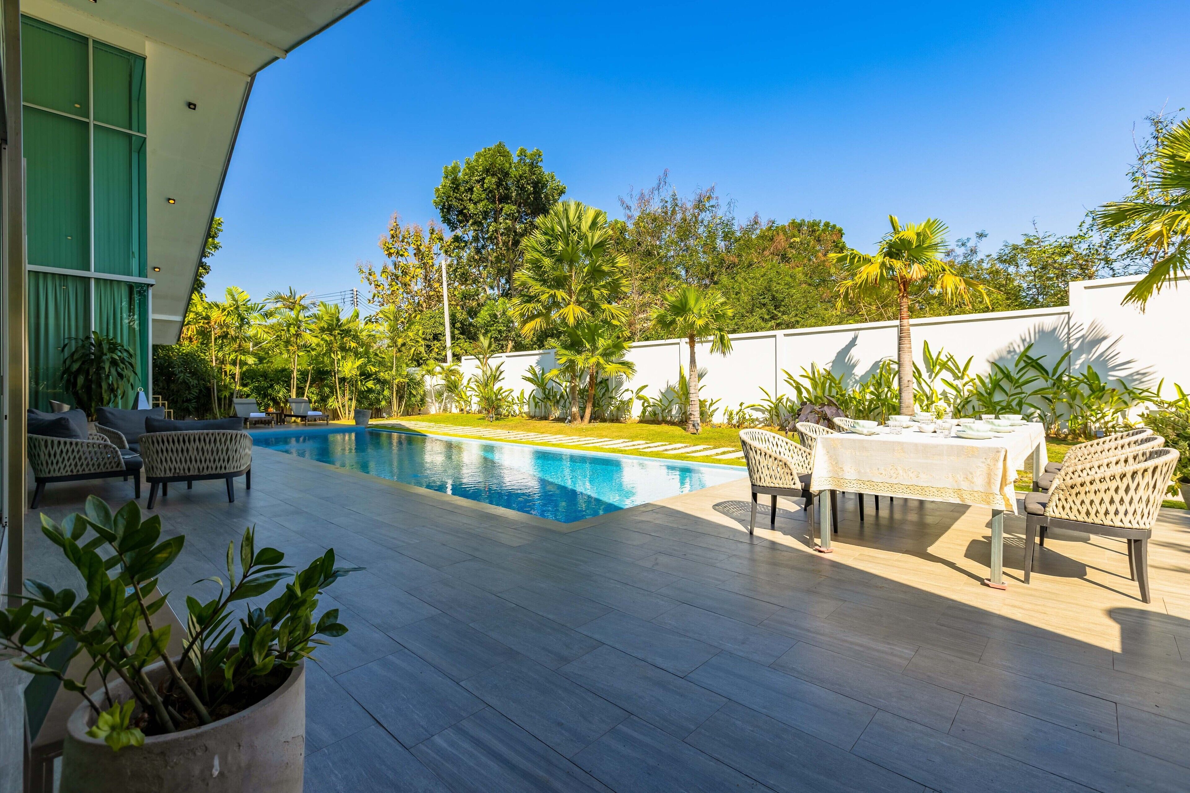 A modern outdoor pool area with a dining table and chairs, surrounded by lush greenery and a clear blue sky.