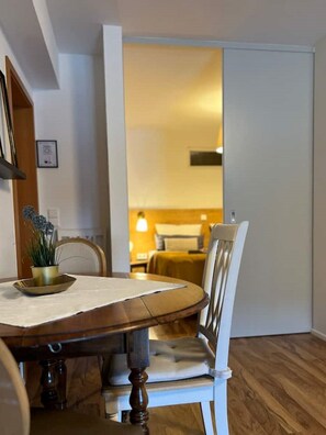 Een eethoek met een houten tafel en stoelen, een klein plantje op tafel, en uitzicht op een slaapkamer met een bed en nachtkastje.