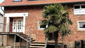 A brick building with a red-tiled roof, a staircase leading to a door marked '1a', and a palm tree in front. - Cozy apartment close to the Rhine (Kehl)