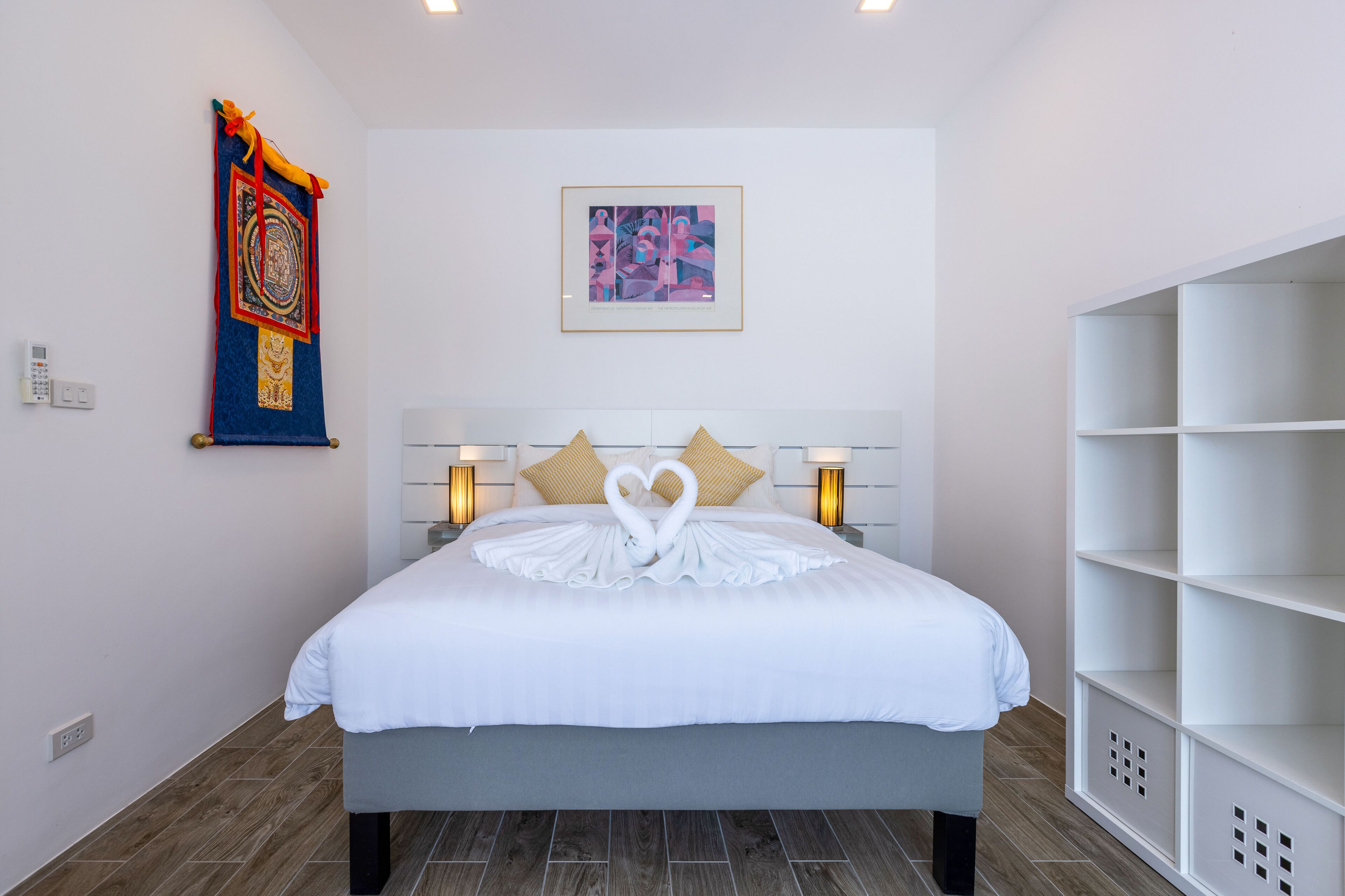 A neatly made bed with a heart-shaped towel arrangement, a colorful tapestry, and a framed abstract painting on the wall.