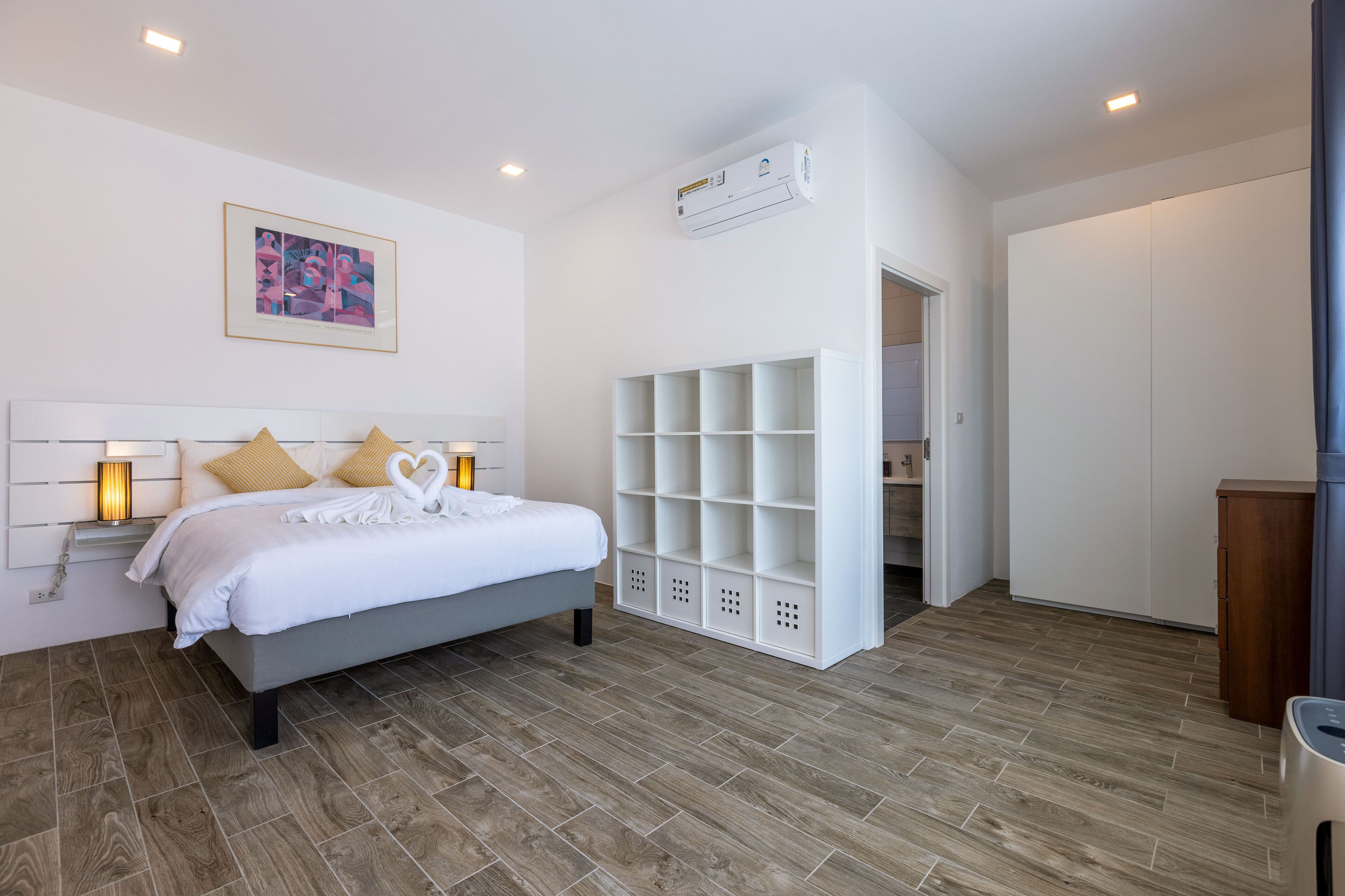 A bedroom with a bed, bedside lamps, a white air conditioning unit, a framed picture, and a built-in shelving unit.