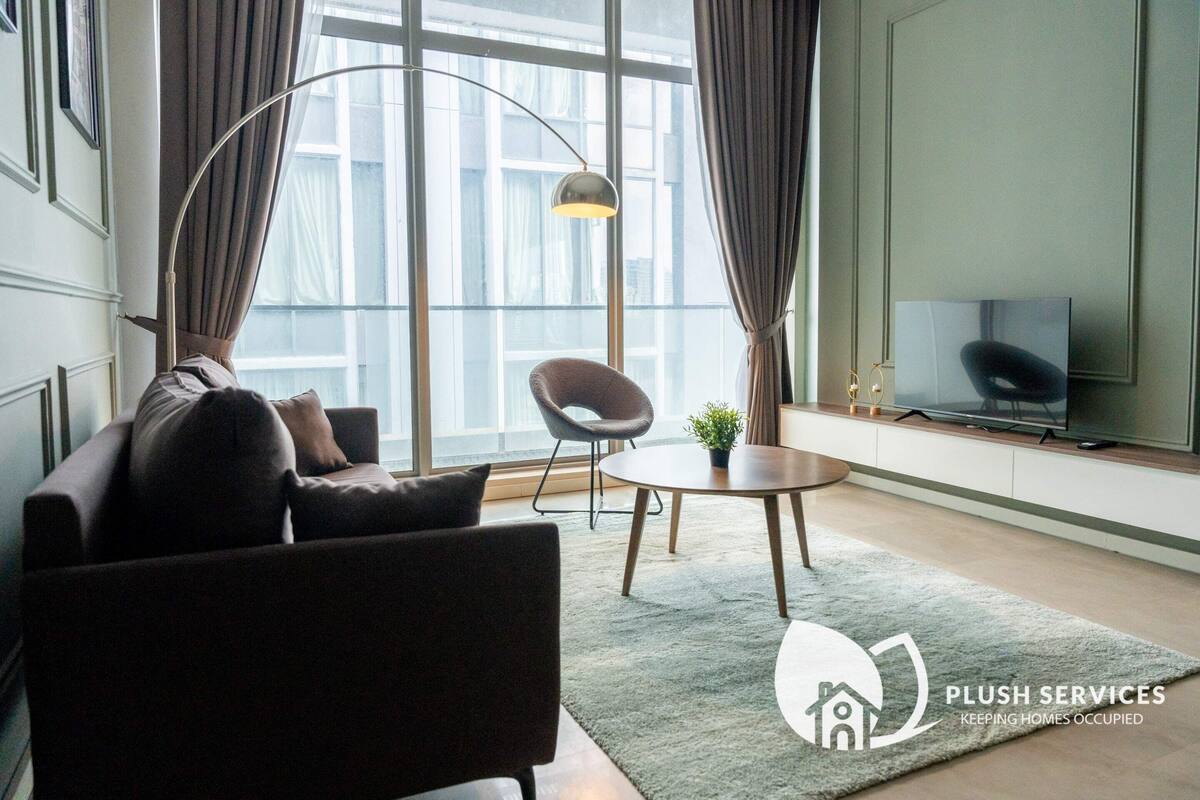 Ein modernes Wohnzimmer mit Sofa, Sessel und einem kleinen Tisch mit einer Pflanze. Ein Fernseher ist an der Wand montiert, und es gibt ein großes Fenster mit Vorhängen.