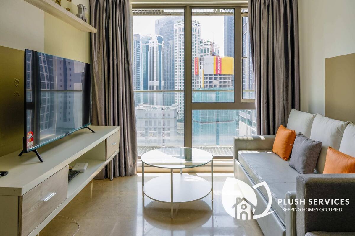 Ein modernes Wohnzimmer mit Fernseher, einem Glastisch und einer Couch mit Kissen. Ein großes Fenster bietet Blick auf Stadtgebäude.