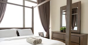 Quarto de hotel com uma cama grande, uma mesinha de cabeceira de madeira, um espelho e uma janela com cortinas.