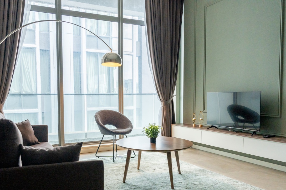 Uma sala de estar moderna com um sofá, poltrona e uma mesinha com uma planta. Uma televisão está instalada na parede, e há uma janela grande com cortinas.