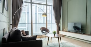 Uma sala de estar moderna com um sofá, poltrona e uma mesinha com uma planta. Uma televisão está instalada na parede, e há uma janela grande com cortinas.