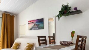 A room with a bed, a dining table with chairs, a painting of a sunset, a mirror, and a shelf with plants. - Bungalow, 10-minute walk from Etang-Salé les Bains beach, sleeps 2 to 4. (L'Étang-Salé)