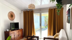 A room with a television on a wooden cabinet, a dining table with chairs, and a sliding glass door leading to an outdoor area with plants. - Bungalow, 10-minute walk from Etang-Salé les Bains beach, sleeps 2 to 4. (L'Étang-Salé)
