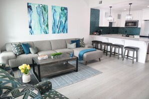 Una sala de estar moderna con un sofá gris, una mesa de centro y una zona de cocina con una isla blanca y taburetes de bar.